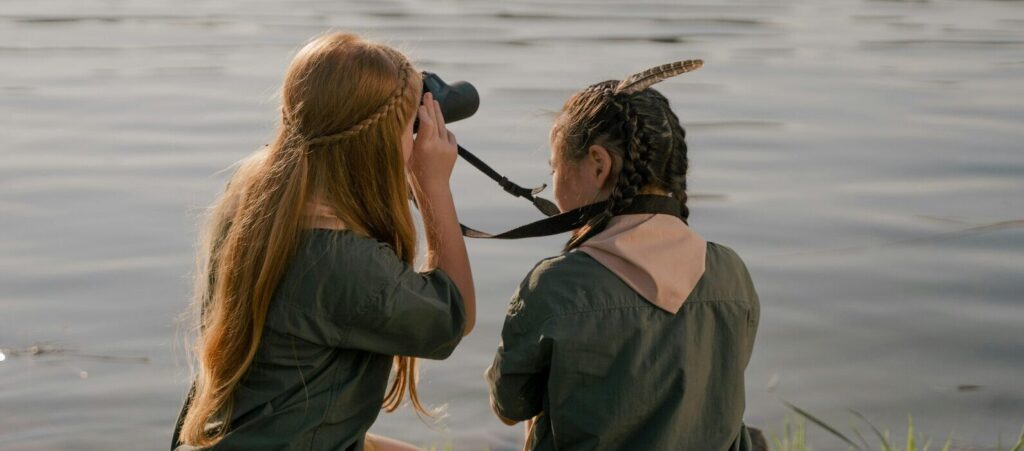 Two girl scouts sit by the water and use binoculars to look out