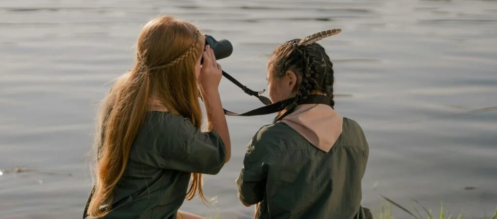 Two girl scouts sit by the water and use binoculars to look out